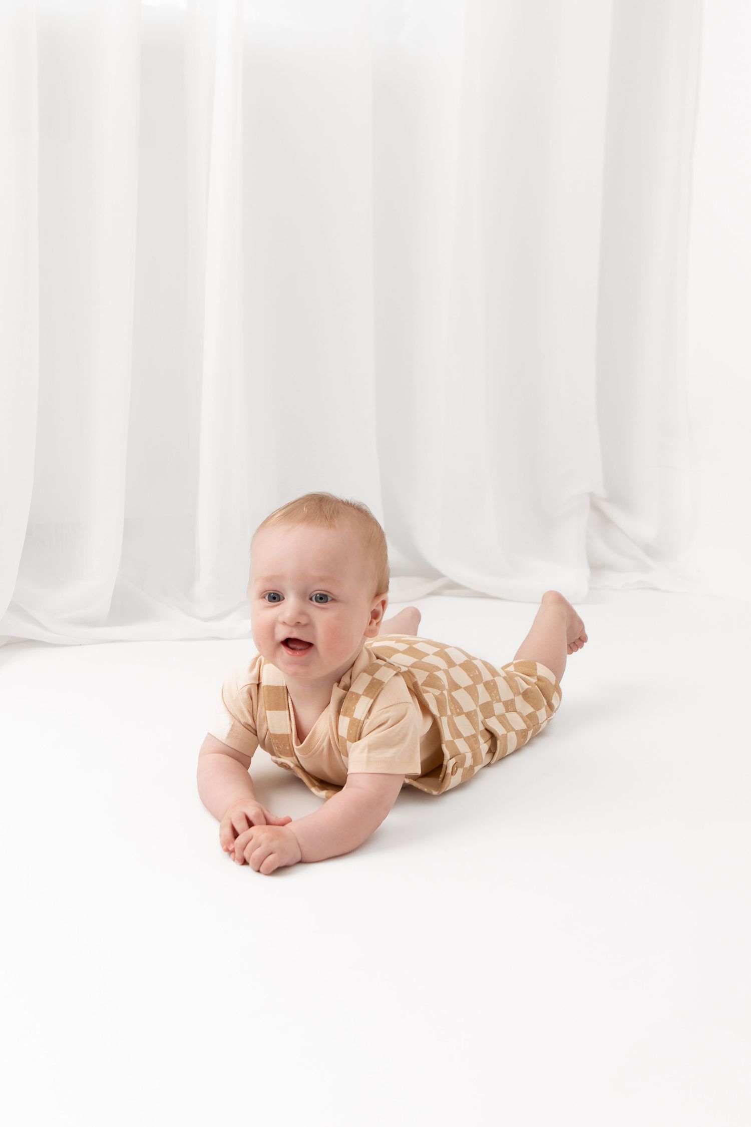 Smiling baby in a beige geometric print romper doing tummy time on a white floor with white curtains.
