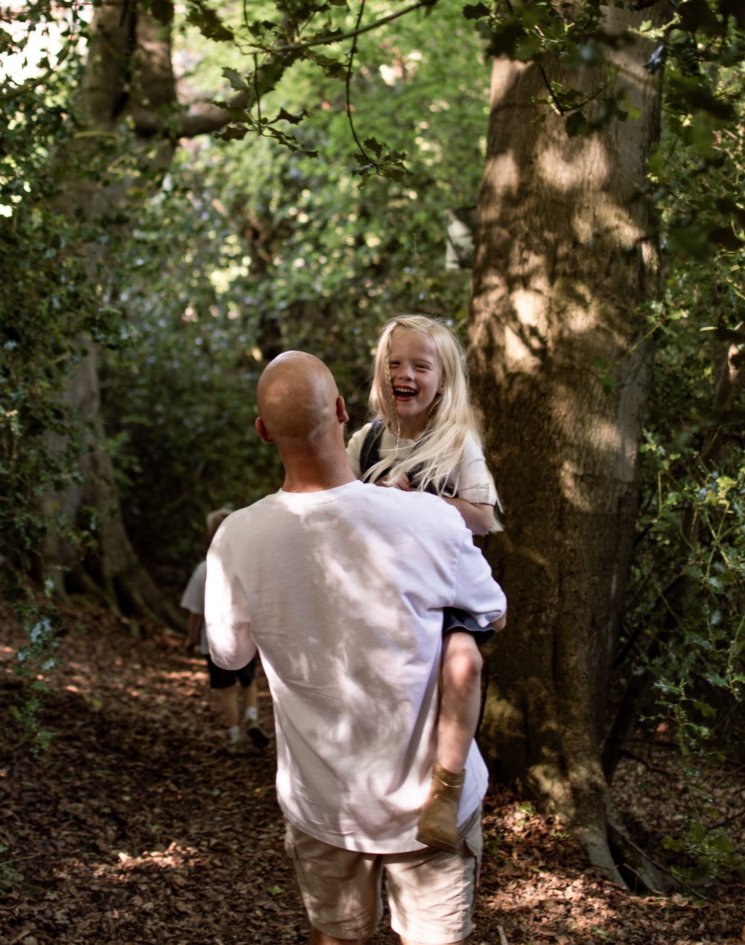 A bald man carries a laughing blonde girl on his shoulders along a sunlit forest path surrounded by green trees.
