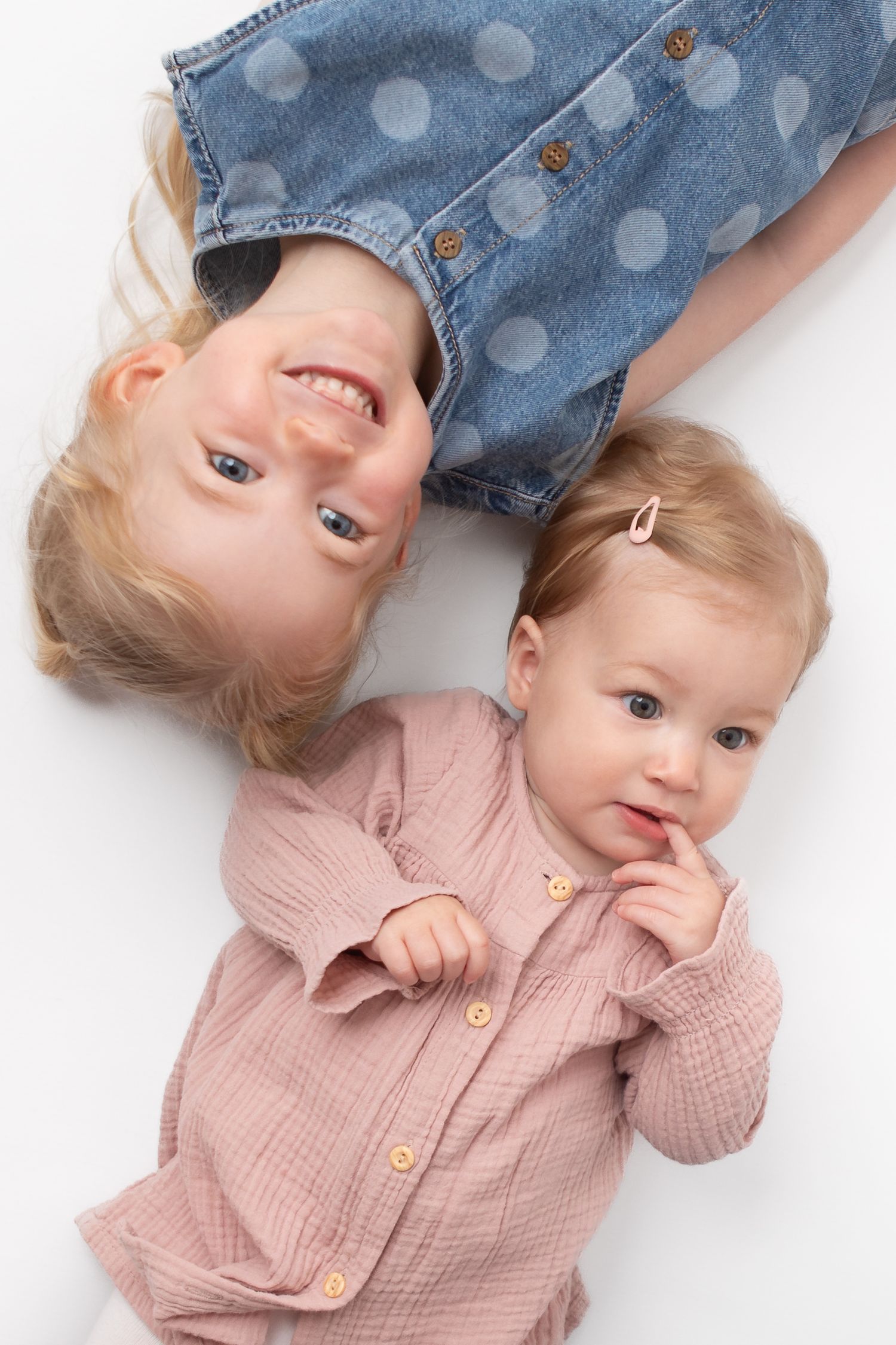 Two young blonde girls lying on white background, older child in denim dress smiling upside down, baby in pink shirt.