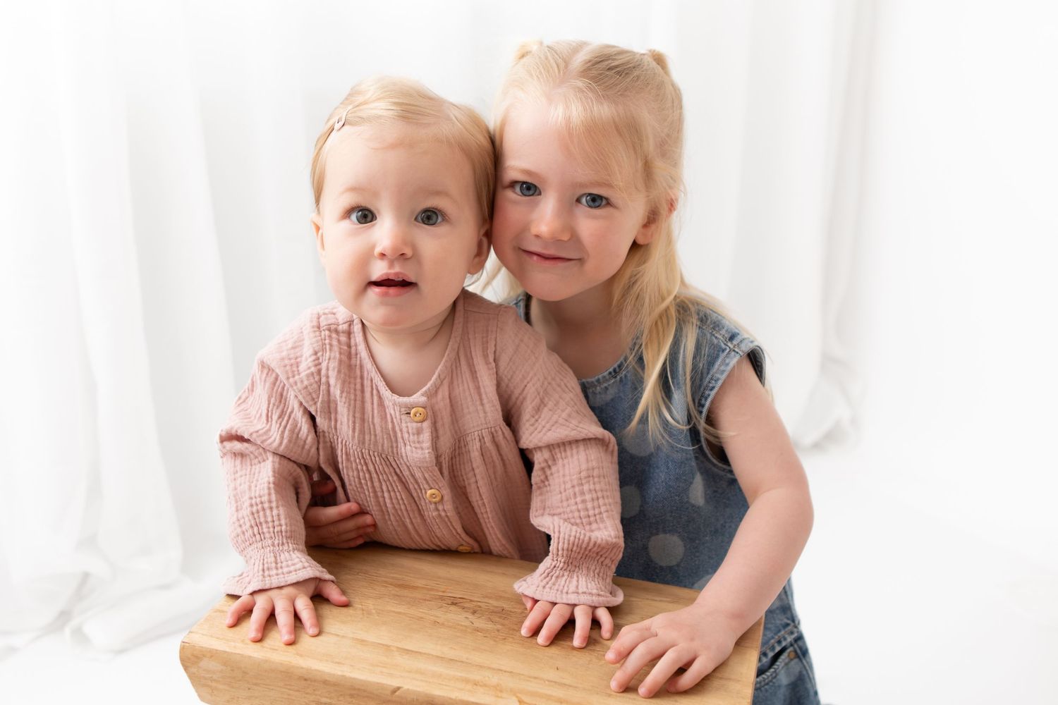 Two young blonde sisters posing together, baby in pink cardigan and toddler in denim dress, white studio background.