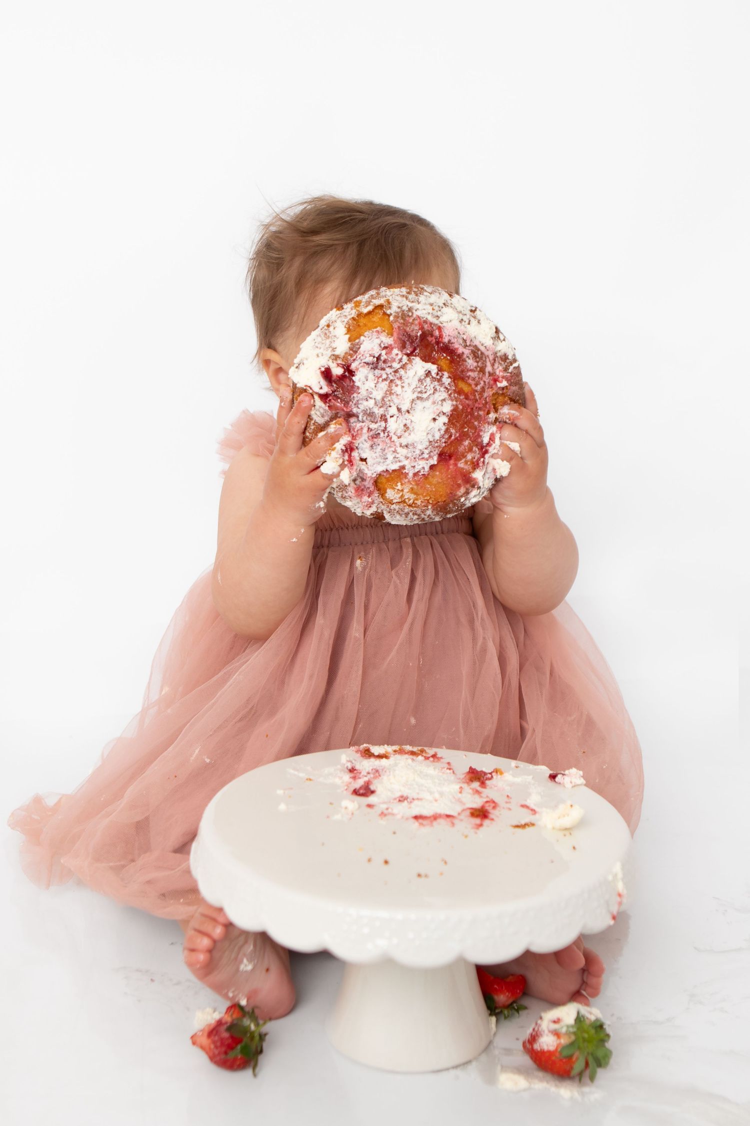 Toddler girl in pink tulle dress holding smashed birthday cake in front of her face during cake smash photoshoot.