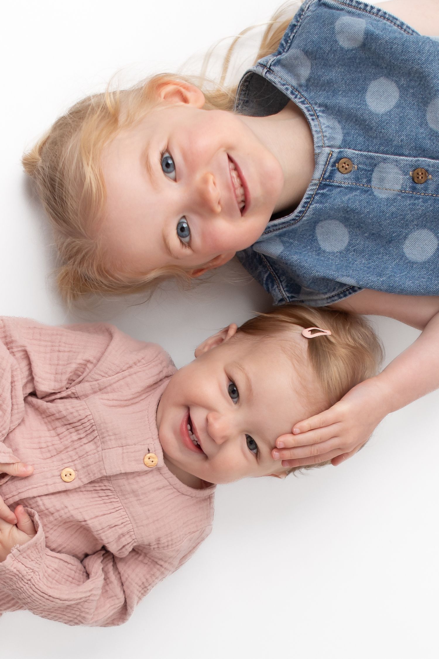 Two smiling blonde toddler girls lying on white background, older sister in denim dress, younger in pink top.