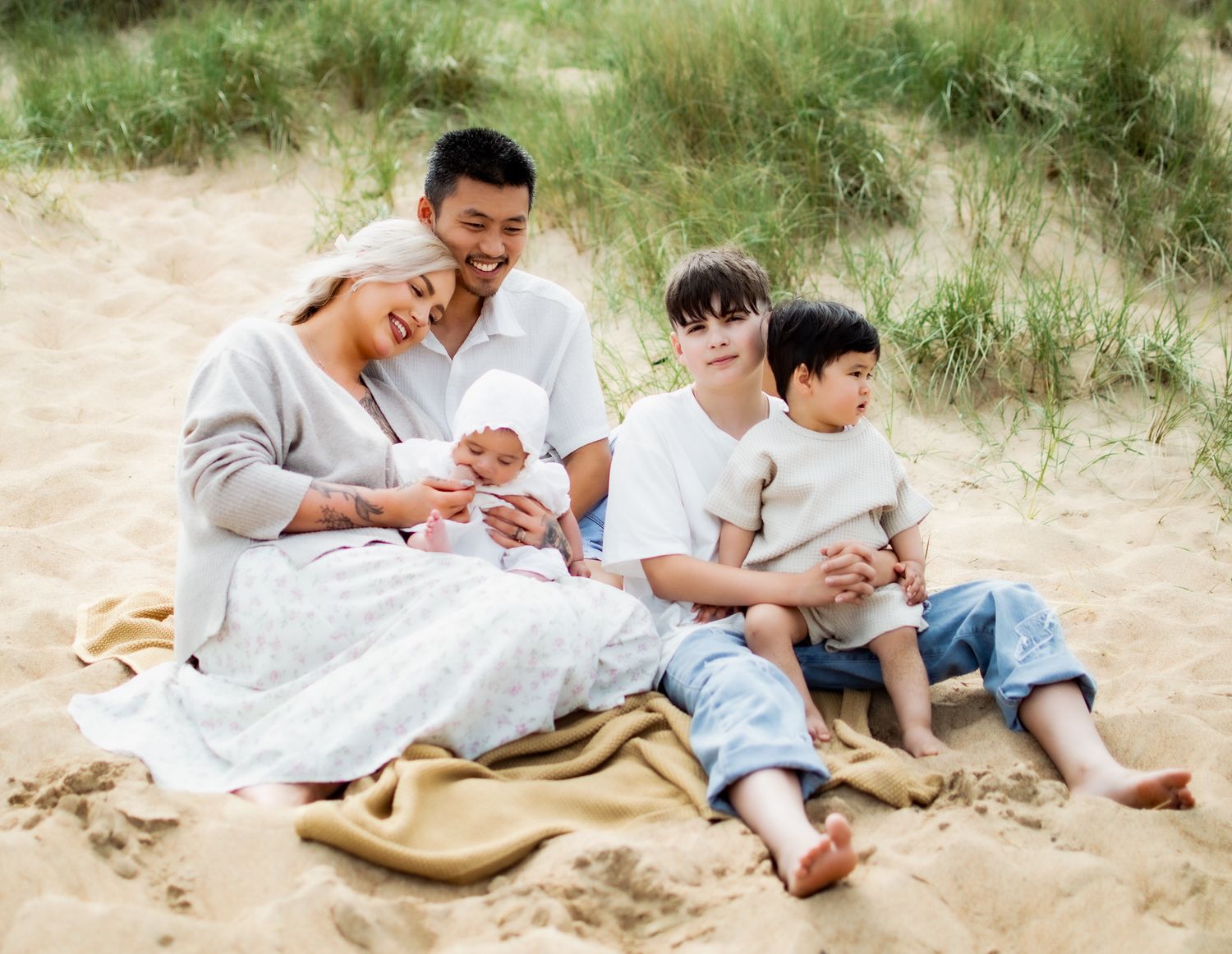 Family of five sitting on sandy beach dunes, parents with newborn baby and two young boys on a blanket.