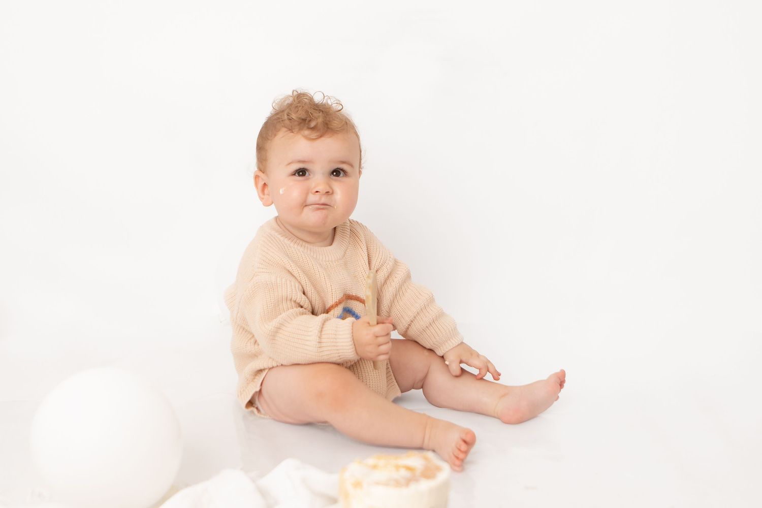 A cute baby in a beige romper sits on a white floor, holding a spoon near a smash cake for their first birthday.
