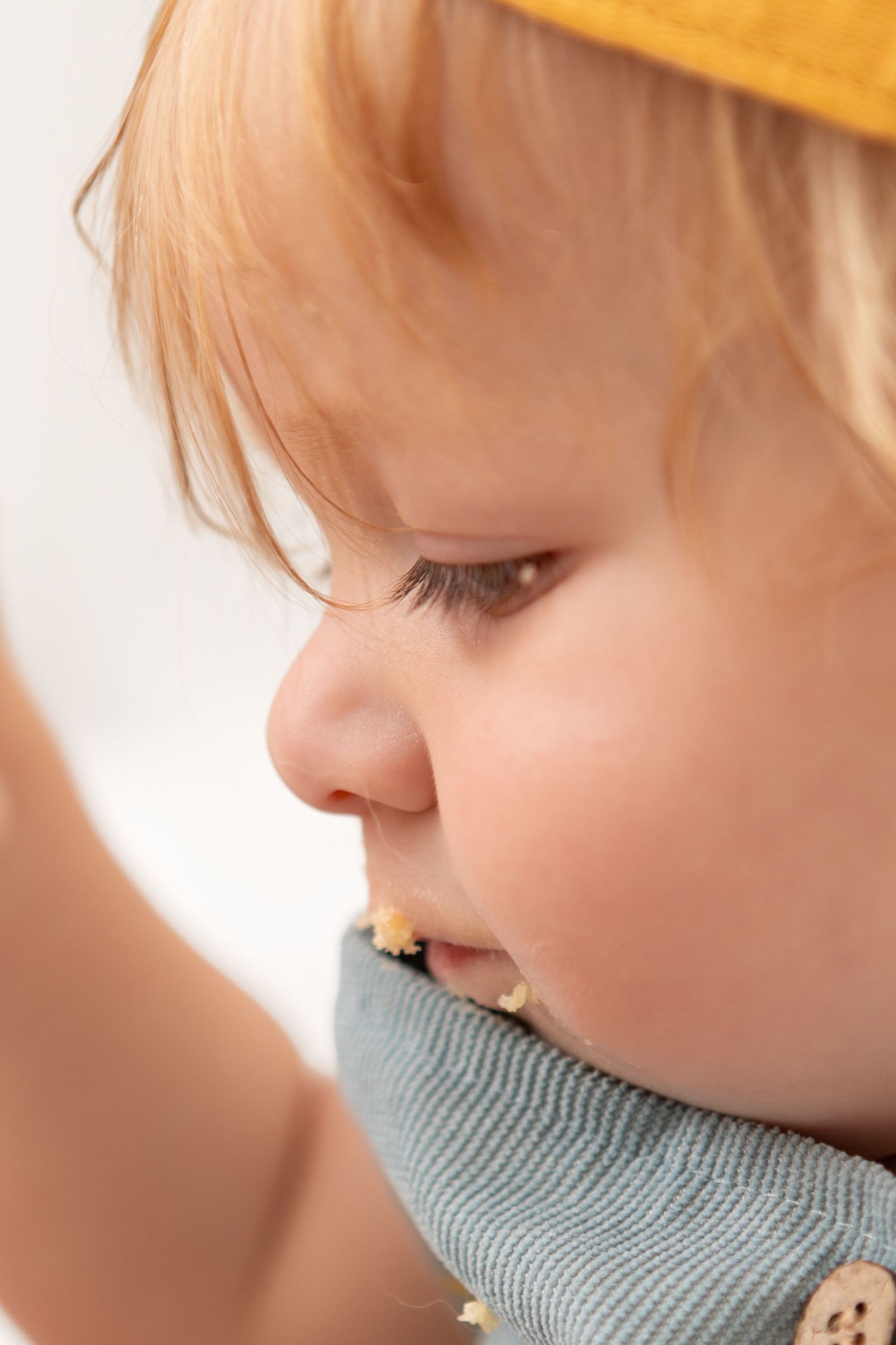 Close-up portrait of a cute baby with red hair wearing a yellow hat and blue bib, eating a snack.