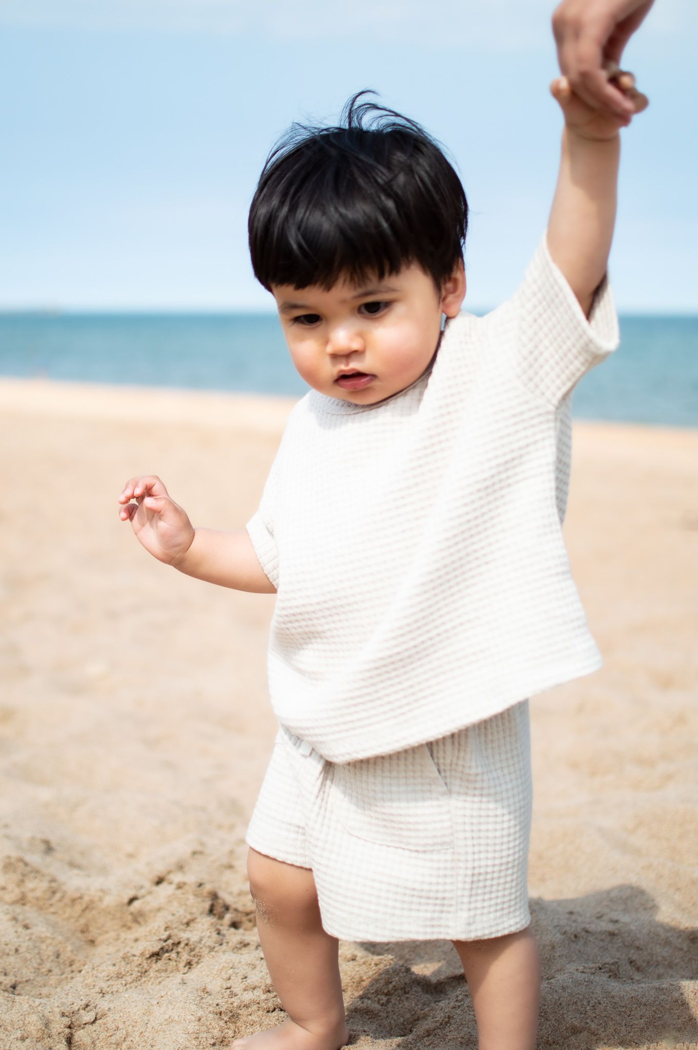 Adorable toddler in white waffle outfit stands on sandy beach holding an adult hand by the ocean.