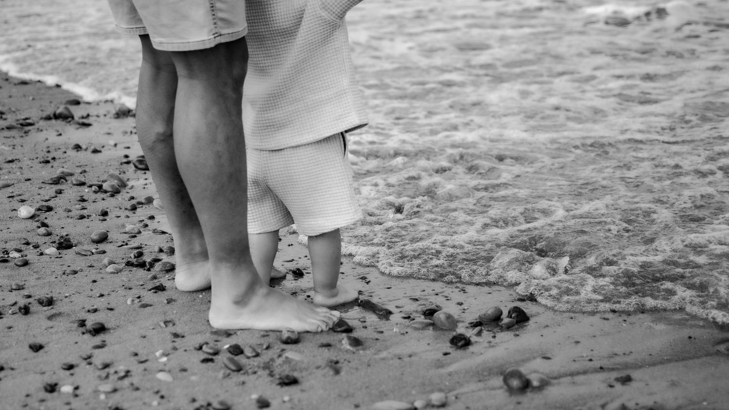Black and white photo of adult and child standing barefoot together on a sandy beach with gentle waves.