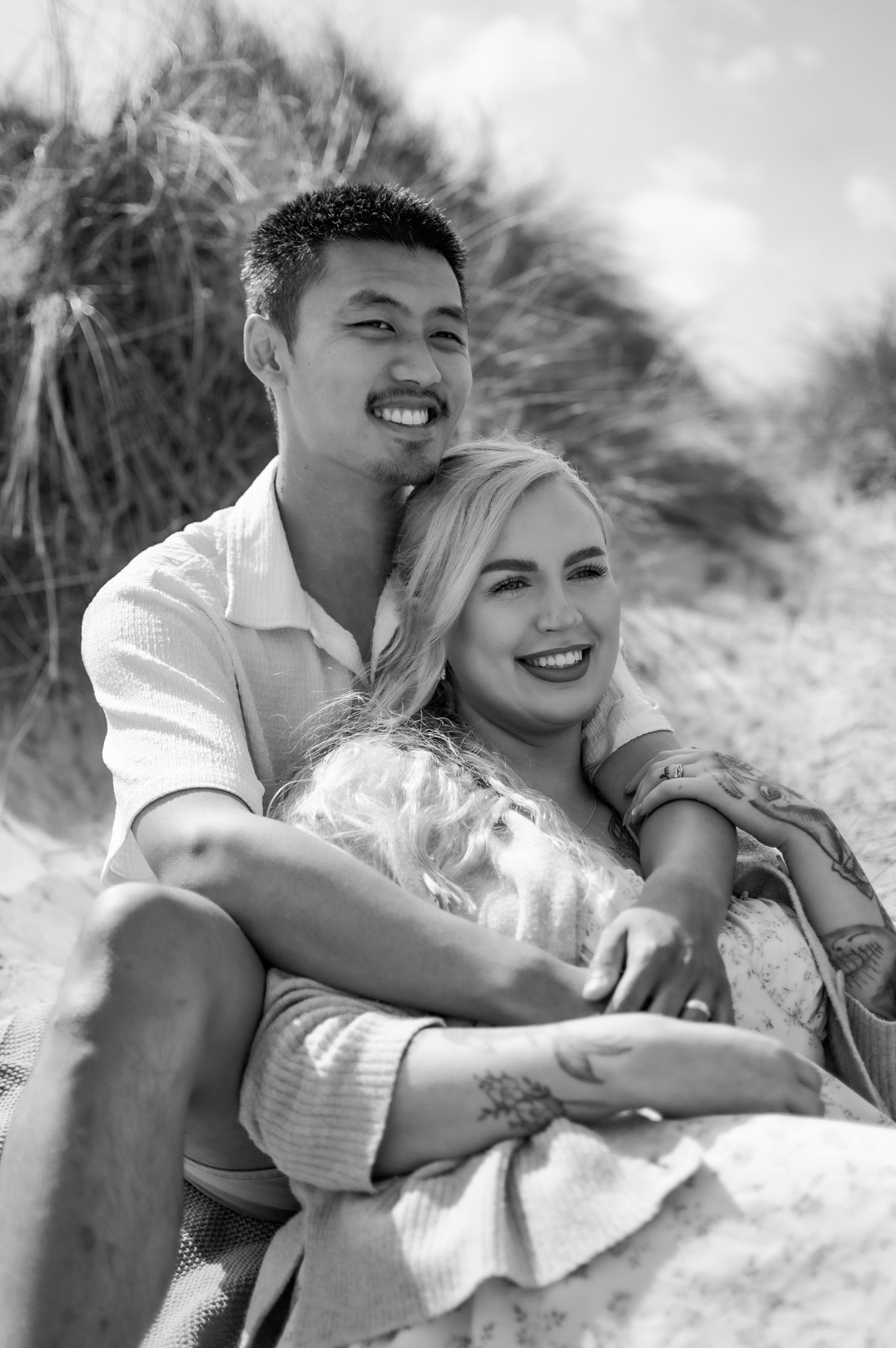 Black and white photo of a smiling couple embracing on sandy beach dunes with grass in the background.