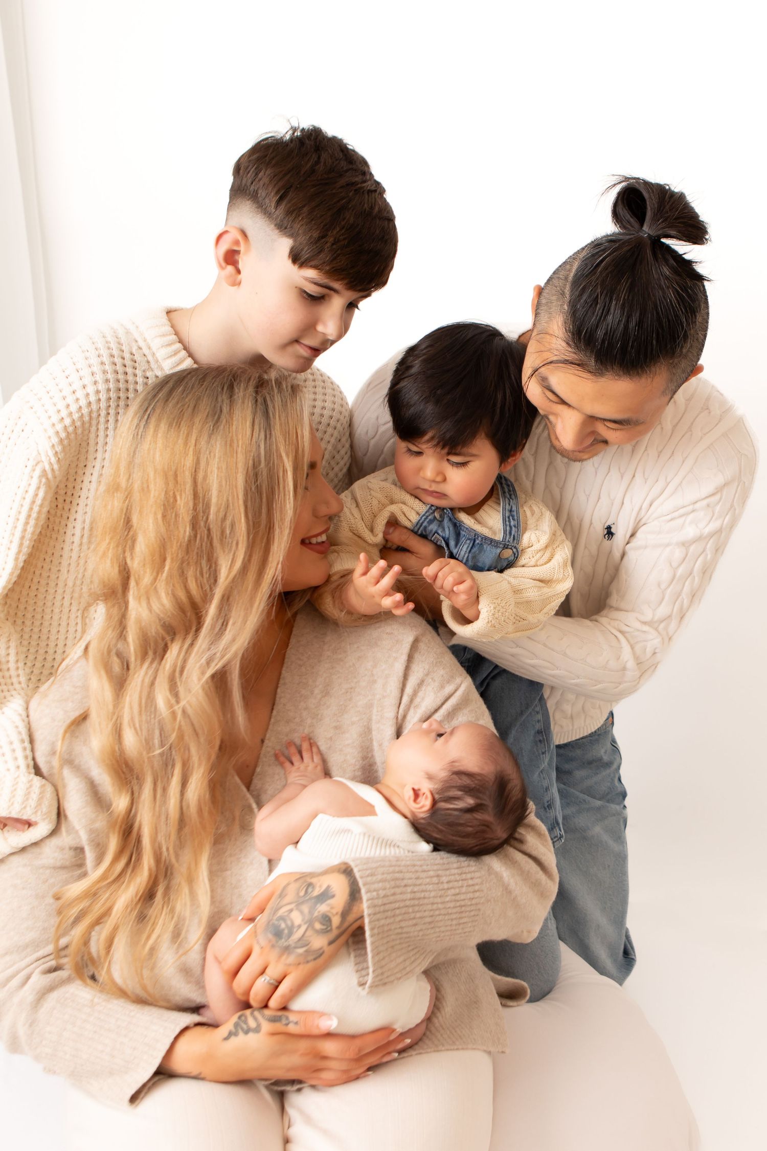 Family gathered around newborn baby, parents and children sharing a tender moment in matching cream sweaters.
