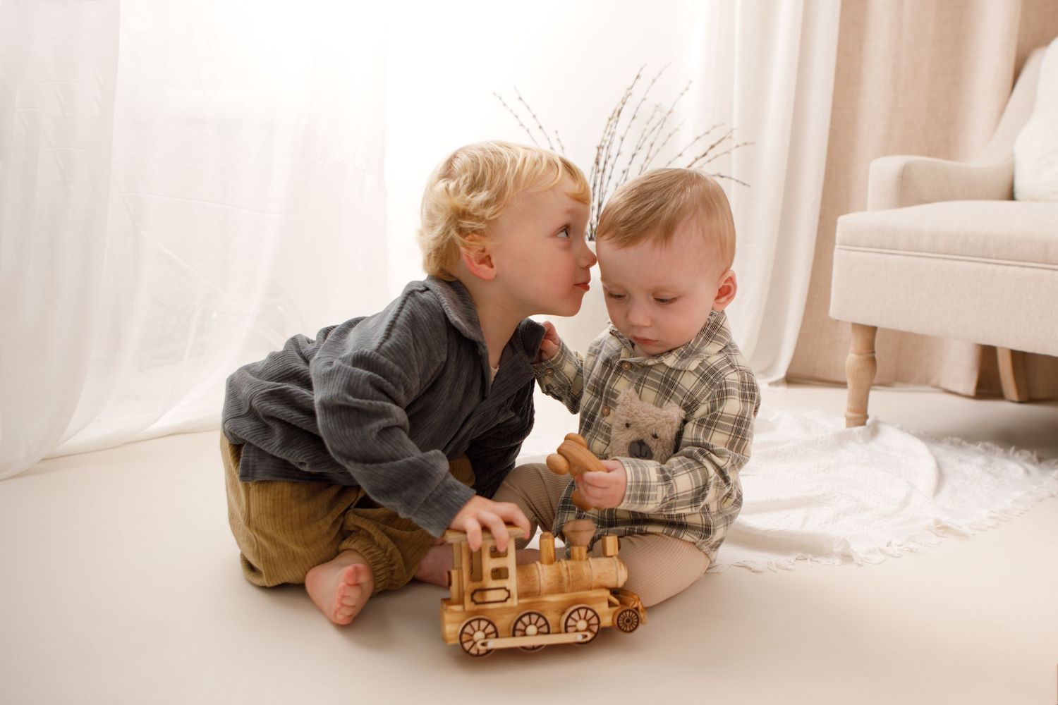 Two toddlers sit on a white floor, sharing a wooden toy train in a bright, cozy room.