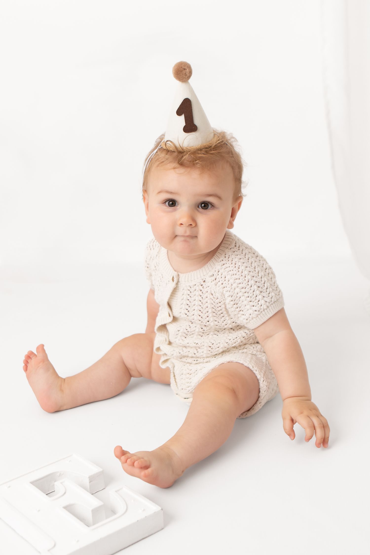 Adorable baby in knit romper wearing a number one birthday hat sits on white background near cake smash props.