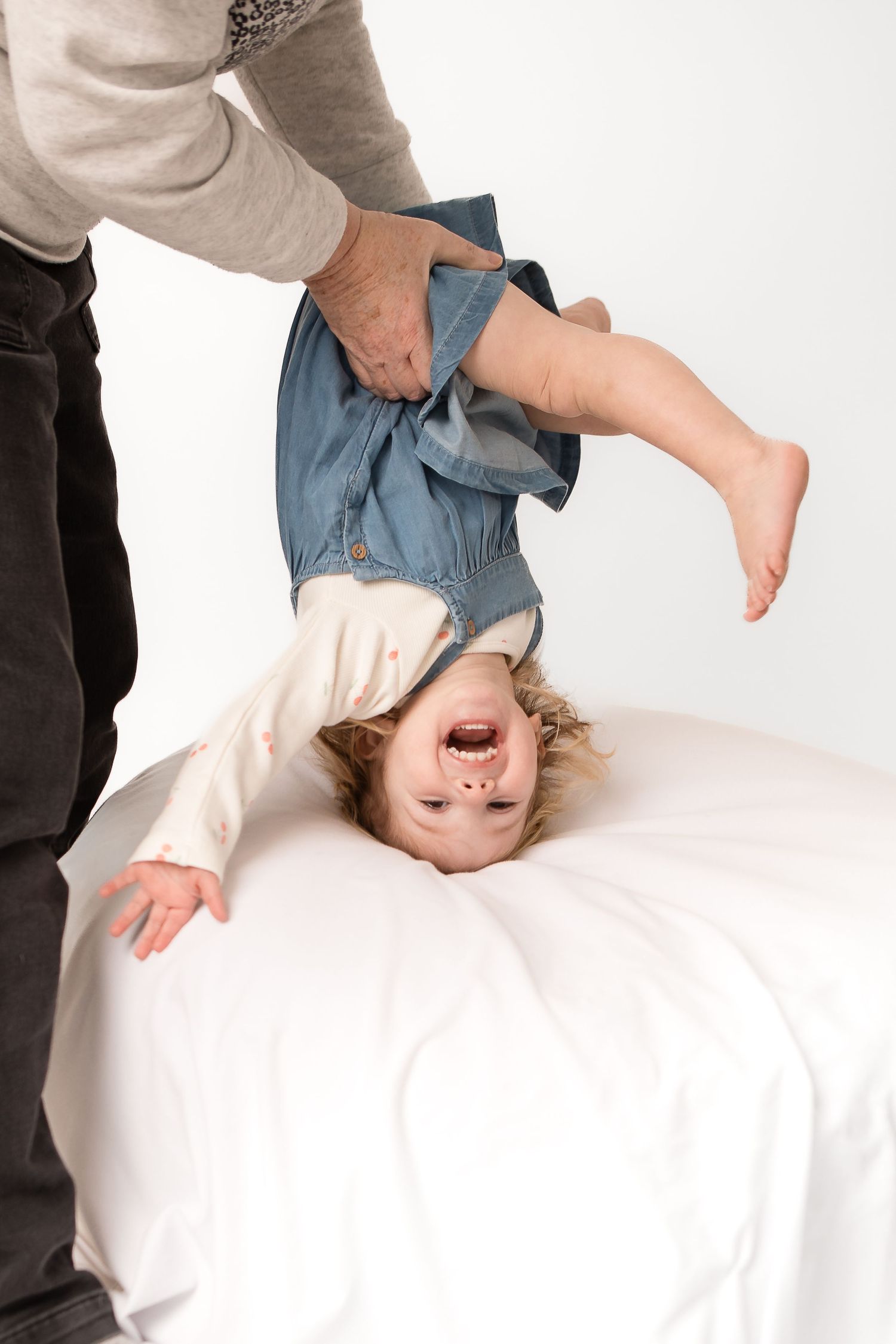 Adult holding a laughing toddler upside down over a white bed, child wearing denim jacket and white outfit.
