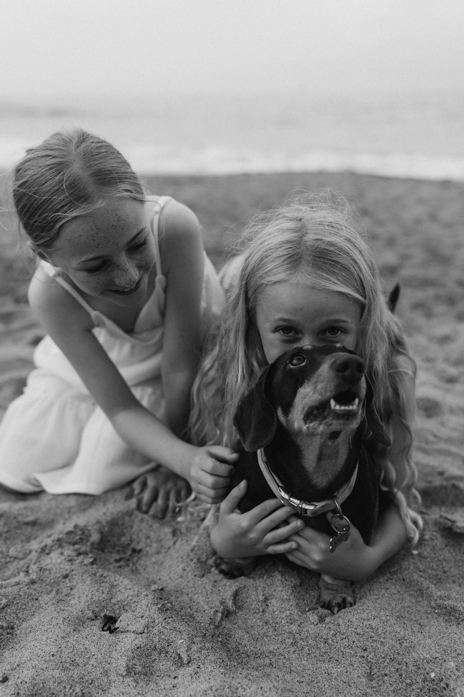 Children laying in sand playing with friendly black dog on beach.