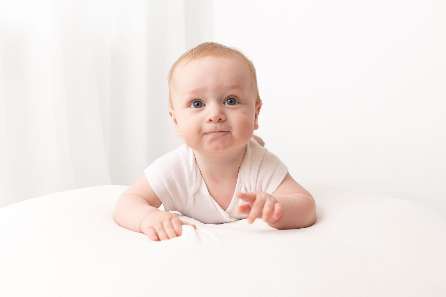 Adorable baby in white onesie lying on stomach on white surface, looking up with curious expression.