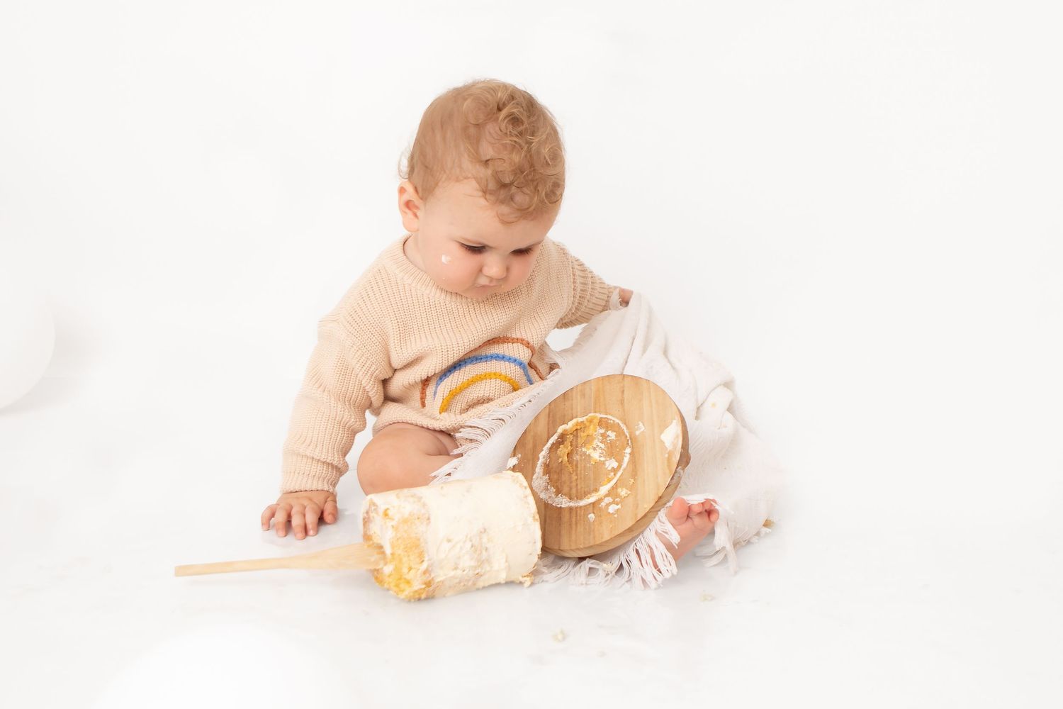Baby in beige sweater sitting on white background, playing with a smashed birthday cake and wooden spoon.