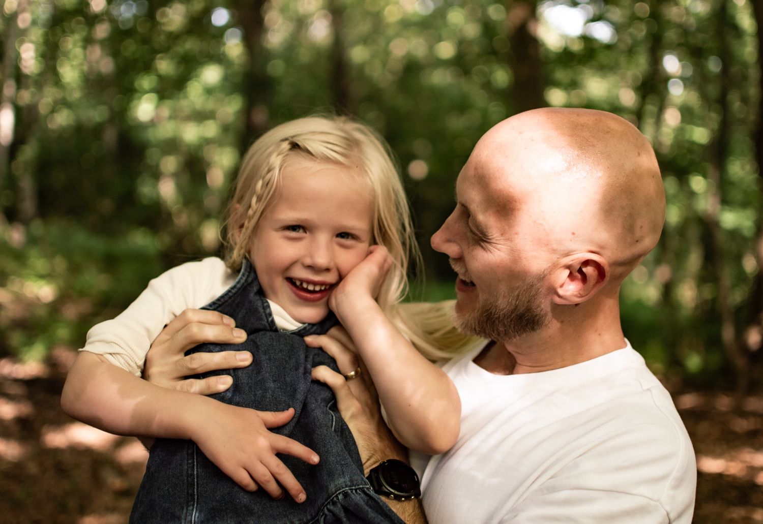 A bald father lovingly holds his laughing blonde daughter in a sunlit forest setting.