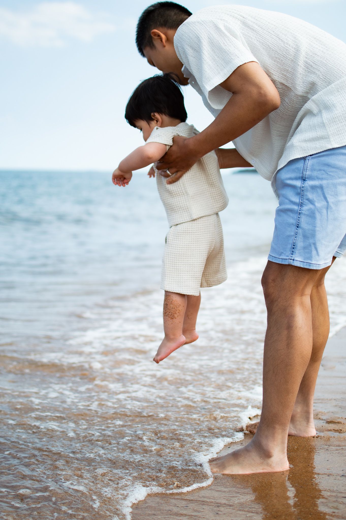 Father lifting toddler at beach shoreline, waves washing over sandy shore on sunny day.
