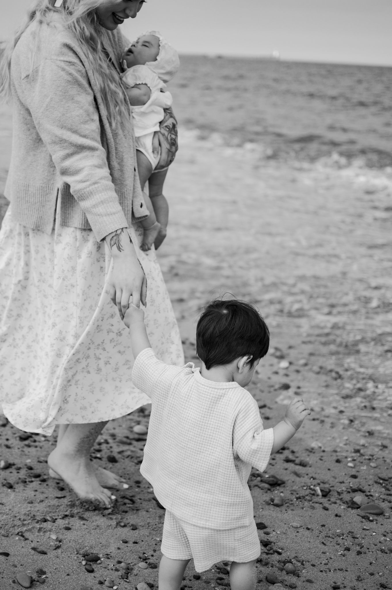 Black and white photo of a mother holding a baby while a toddler walks beside her on a rocky beach.