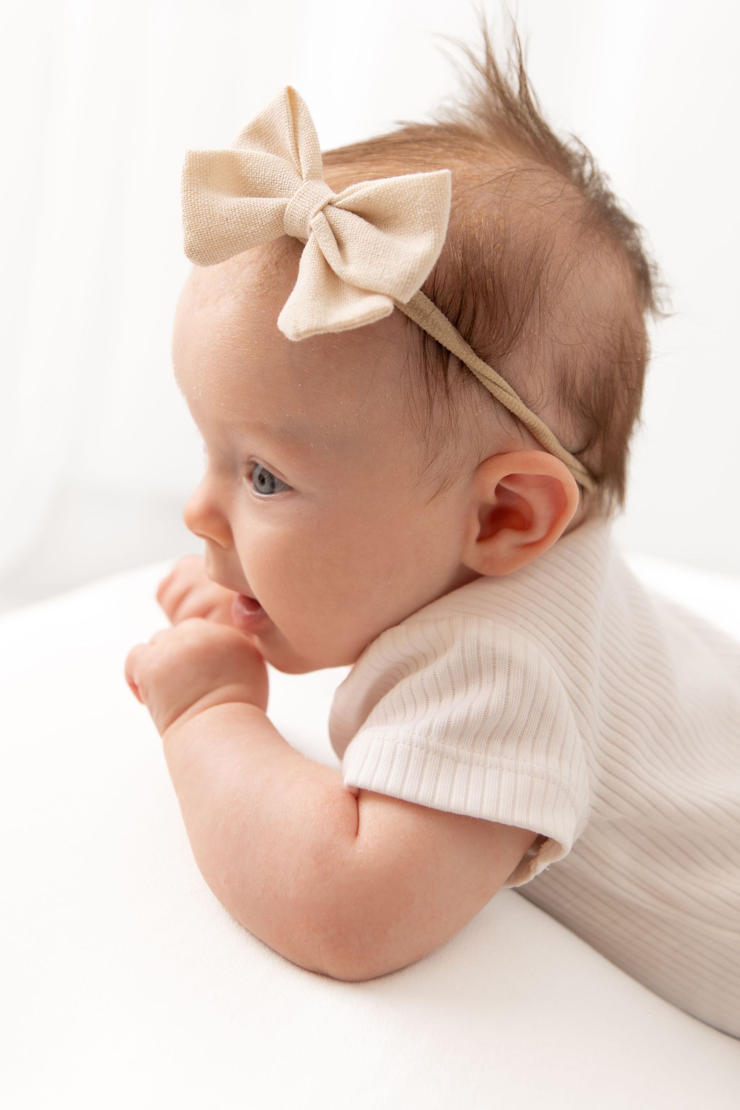 Adorable baby girl wearing a beige bow headband and ribbed onesie, lying on her tummy with fingers in her mouth.