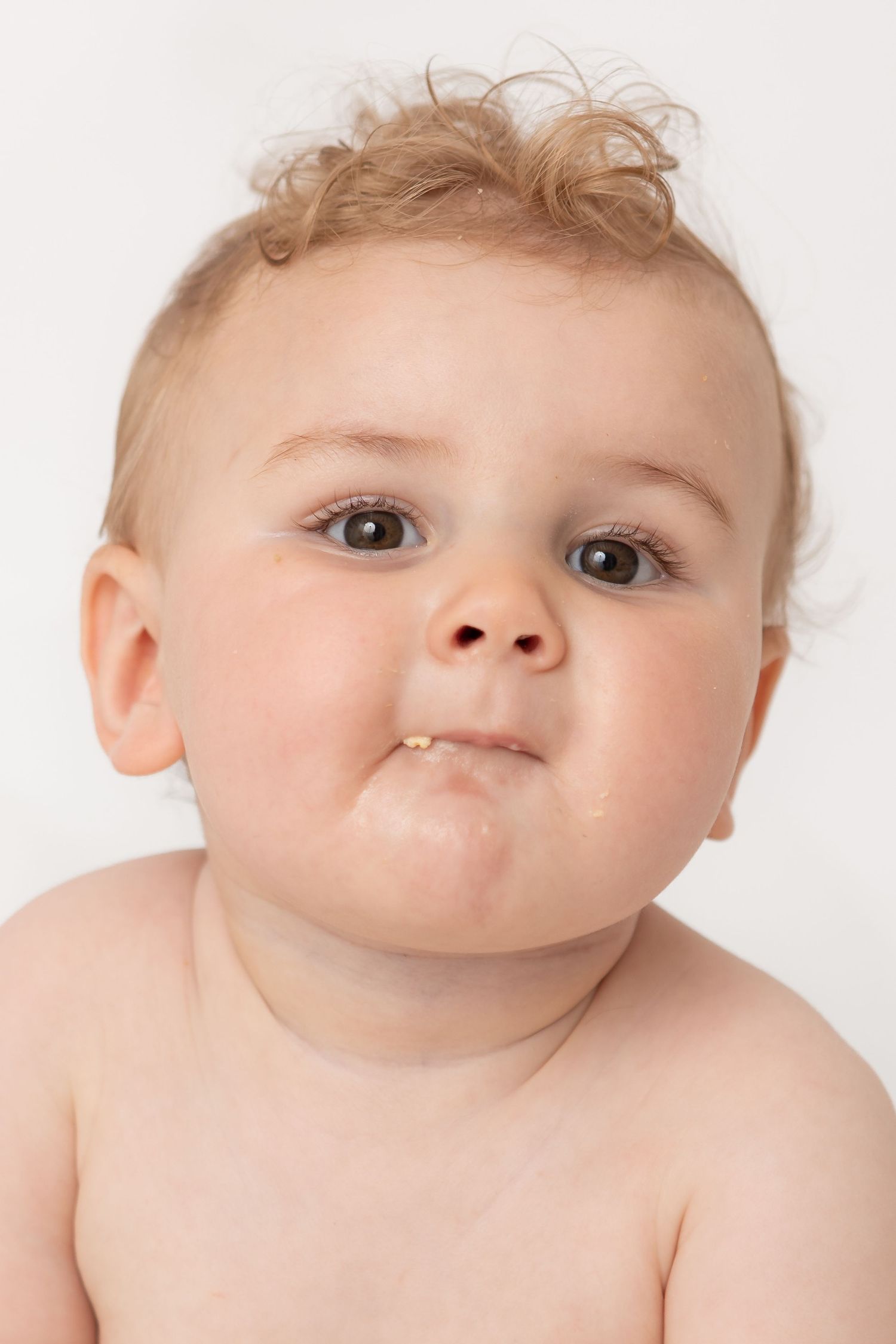 Adorable baby with blonde hair and chubby cheeks making a funny face against a white background.