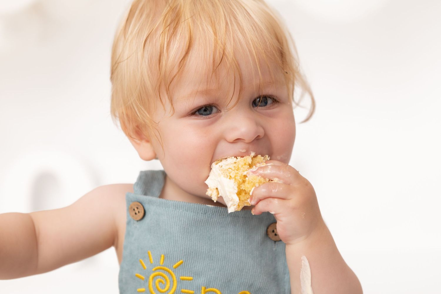 Cute toddler with blonde hair eating a piece of cake, wearing a gray sun-print bib.