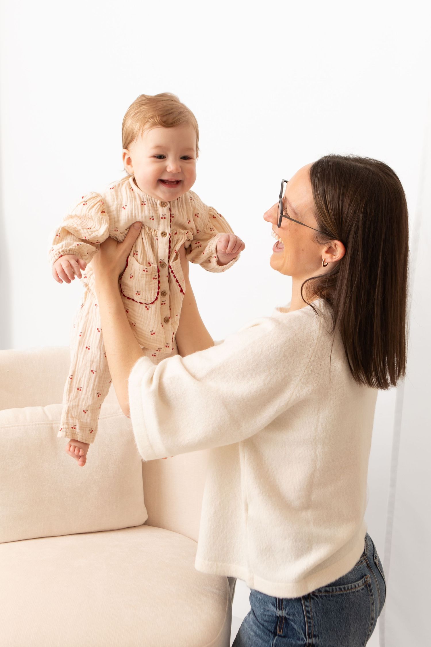 A smiling mother in glasses lifts her happy baby girl in a floral romper above a cream sofa.