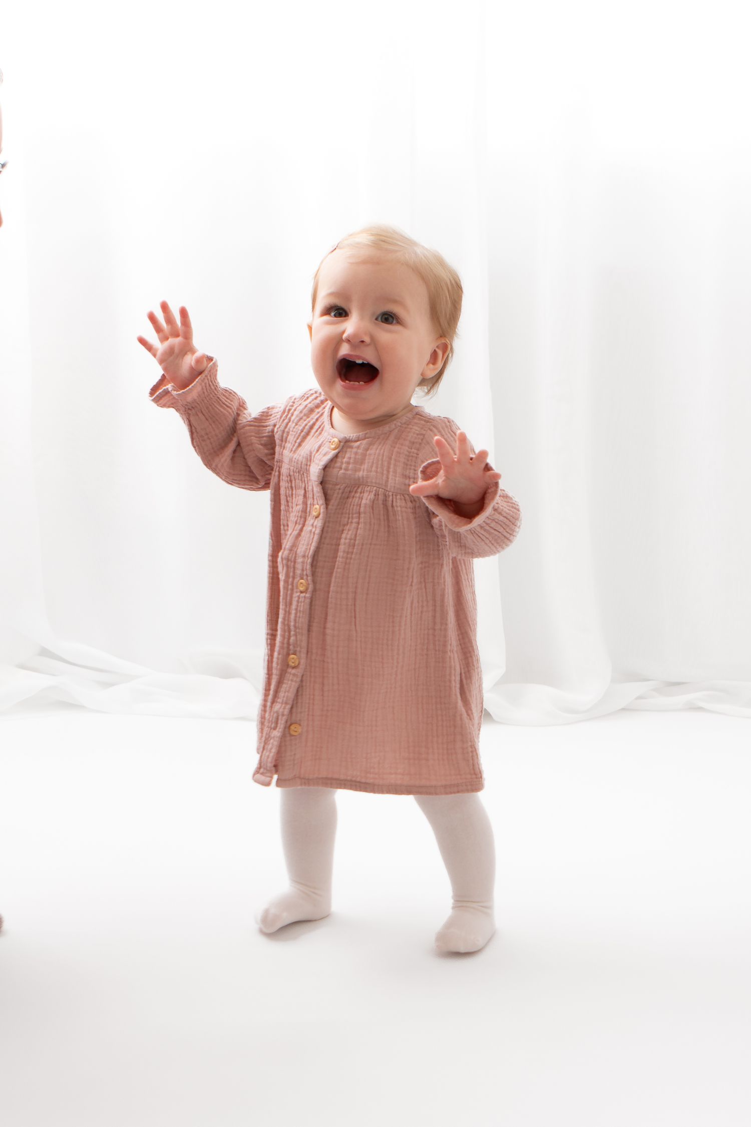 Happy toddler girl in pink muslin dress and white tights standing and laughing with arms raised.