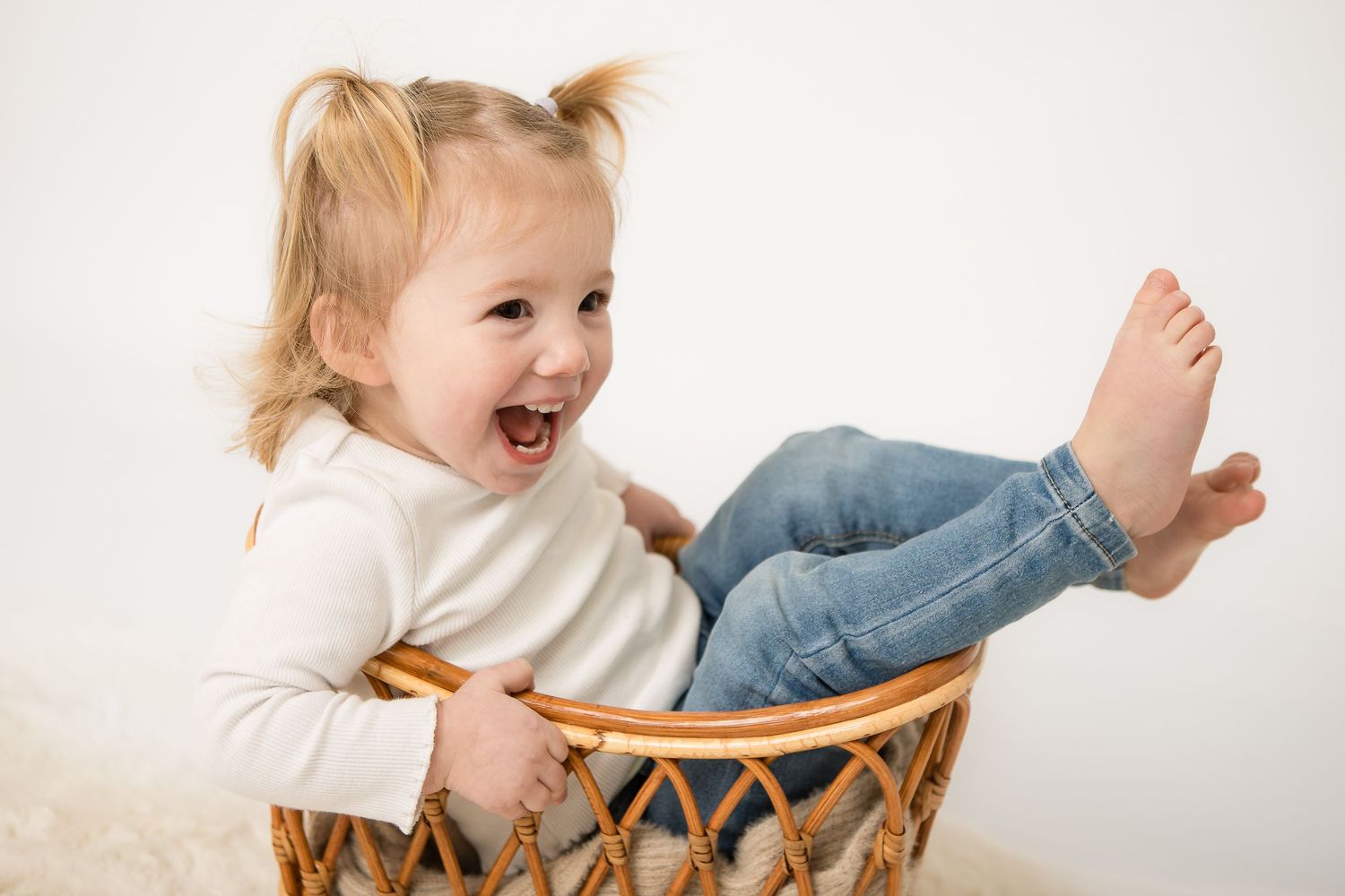 Laughing toddler girl with pigtails sitting inside a wicker basket, wearing jeans and white top.