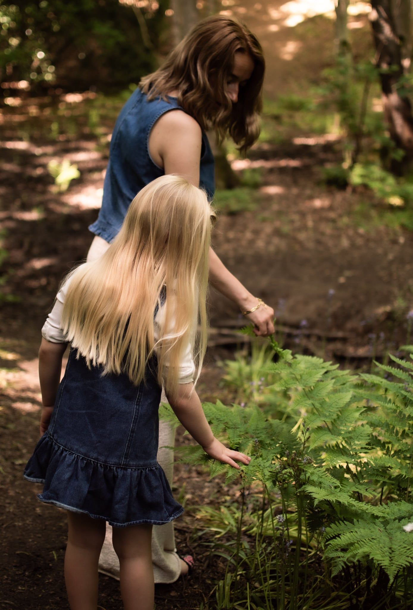 A woman and young blonde girl explore ferns together on a woodland path.