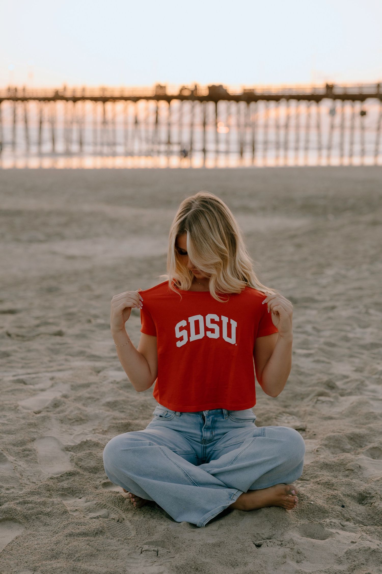 Woman sitting cross-legged on beach sand wearing red SDSU crop top and jeans, with pier in background at sunset.