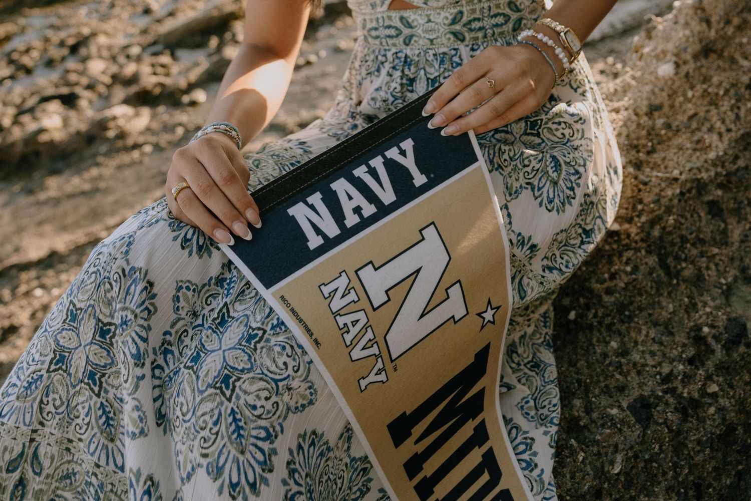 Woman in floral dress holding a Navy pennant flag while sitting on rocks outdoors.