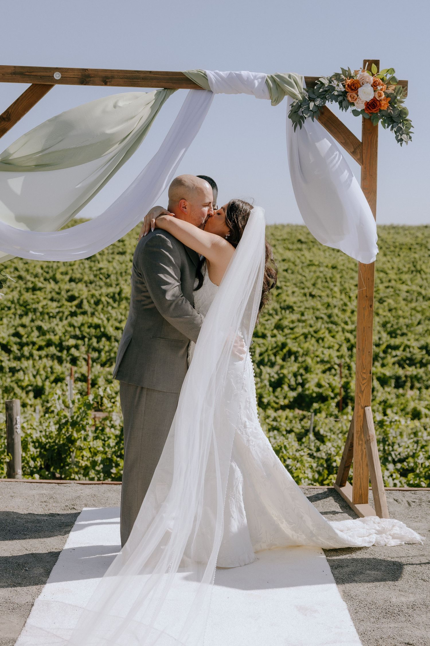 Bride and groom share first kiss under wooden arch with white draping and floral accents at outdoor vineyard wedding ceremony.