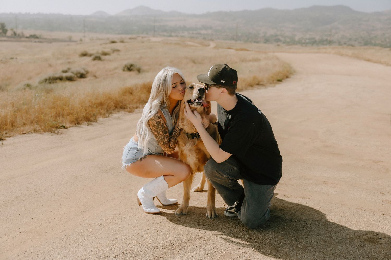 Couple crouching on a dirt road kissing their golden dog with dry hills in the background.