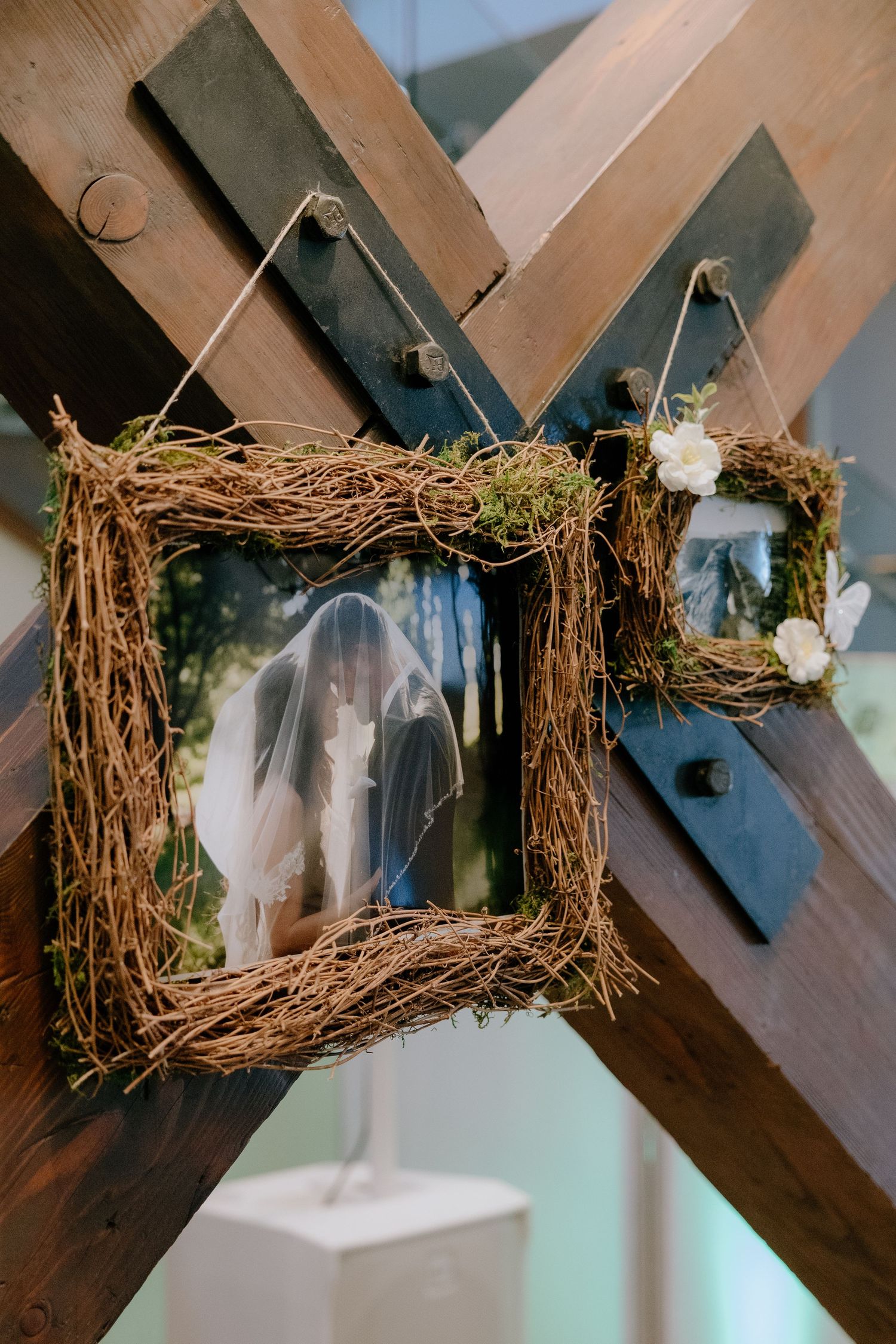 Rustic twig frames with moss and white flowers displaying wedding photos, hanging on wooden beams at a wedding venue.