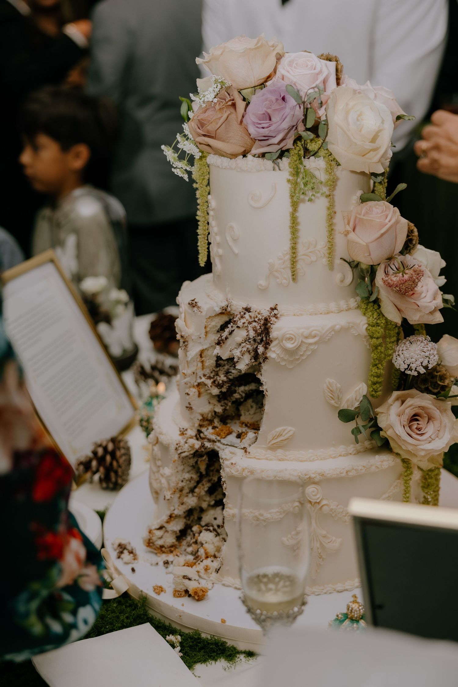 A elegant four-tier white wedding cake adorned with pastel roses and greenery, with a slice removed.