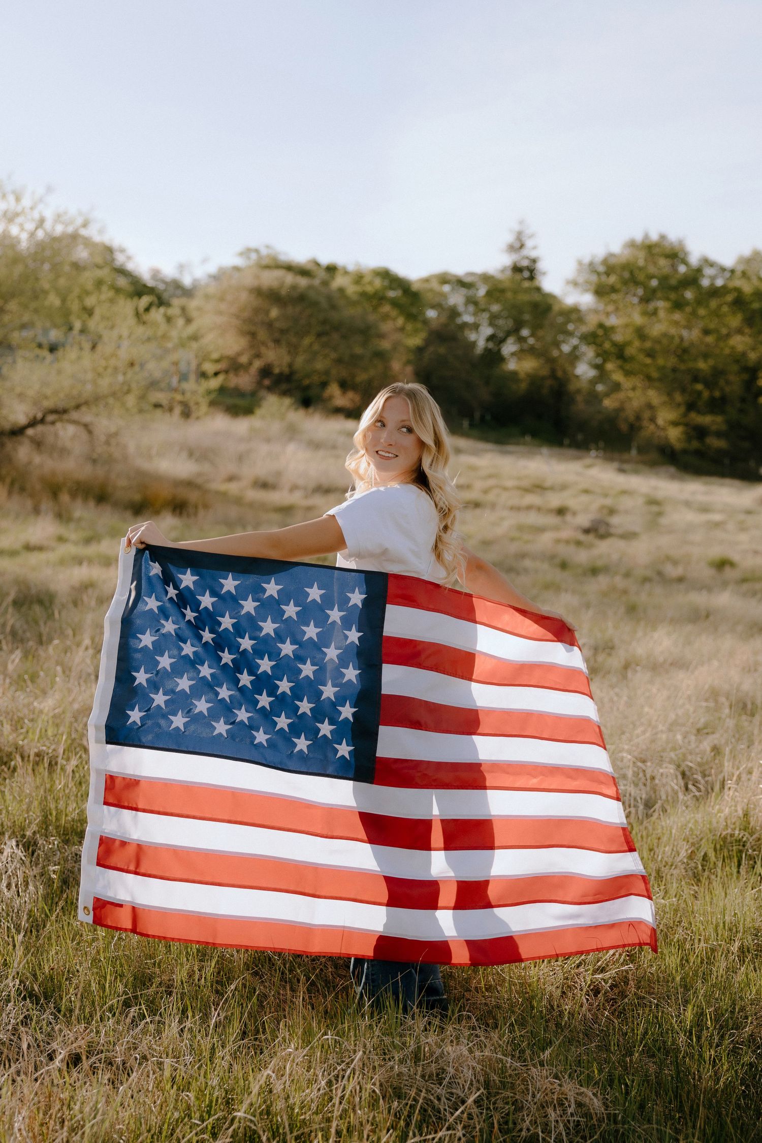Smiling blonde woman holding a large American flag in a sunny green field with trees in the background.