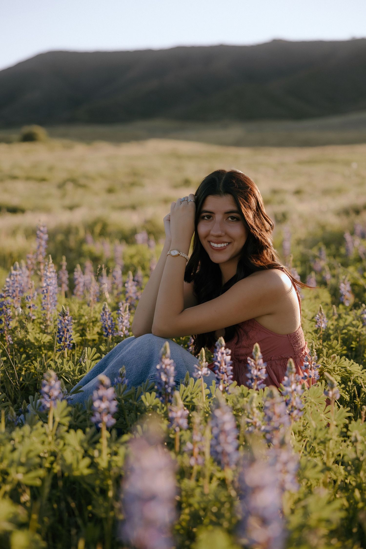Young woman smiling in a field of purple lupine wildflowers at golden hour with rolling hills in background.