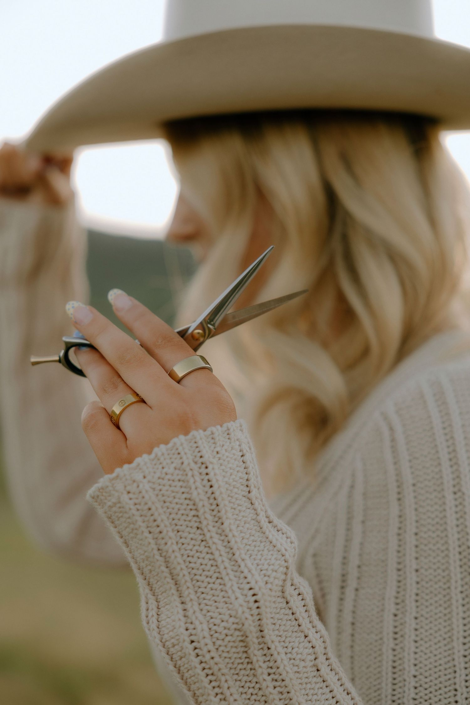 Blonde woman in a cream knit sweater and wide-brim hat holding professional hair scissors outdoors.