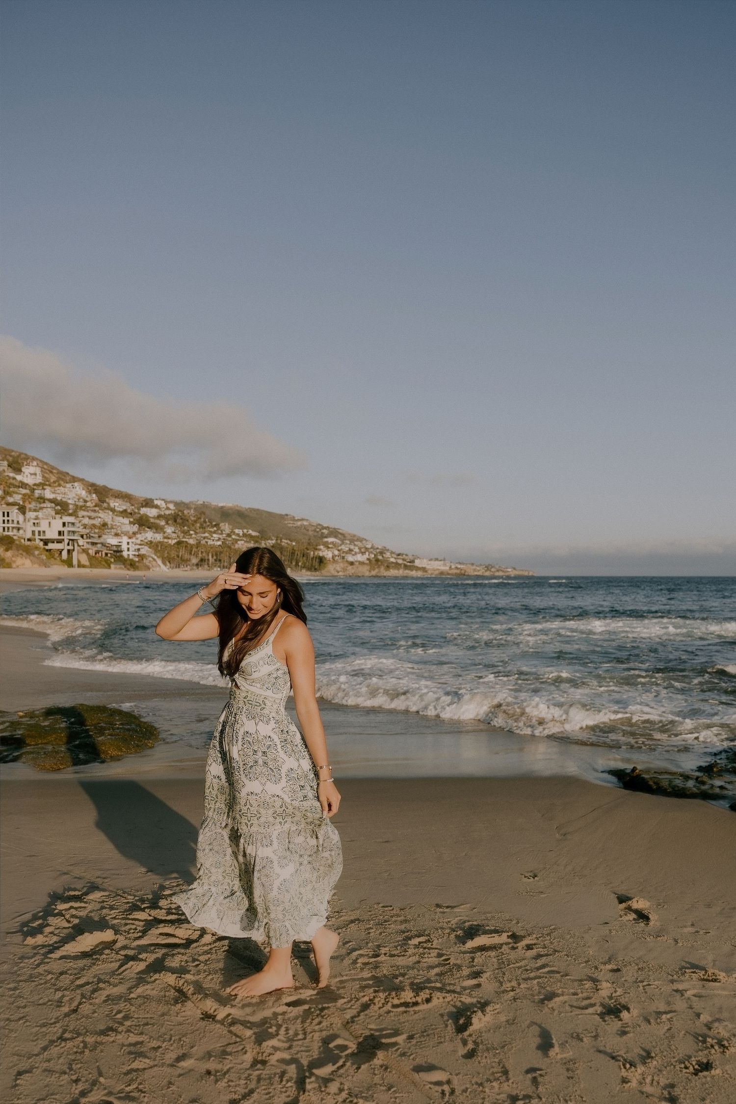 Woman in floral maxi dress walking barefoot on sandy beach with ocean waves and hillside homes in background.