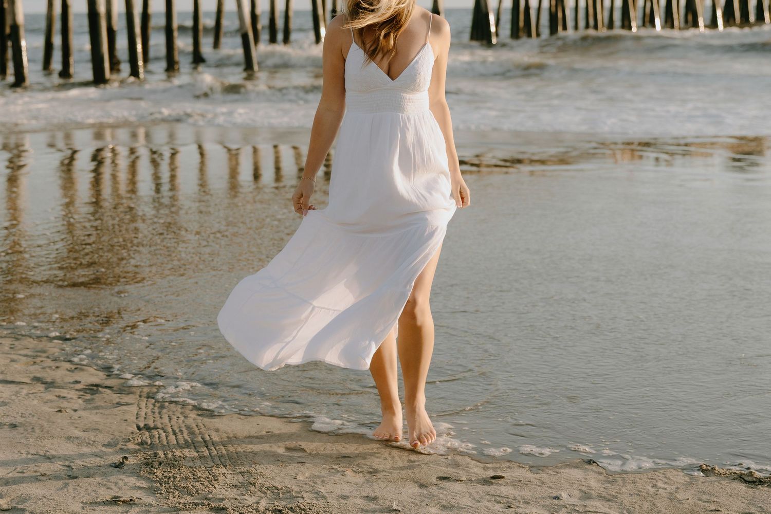 Woman in white flowing dress walking barefoot on sandy beach near ocean waves with wooden pier in background.