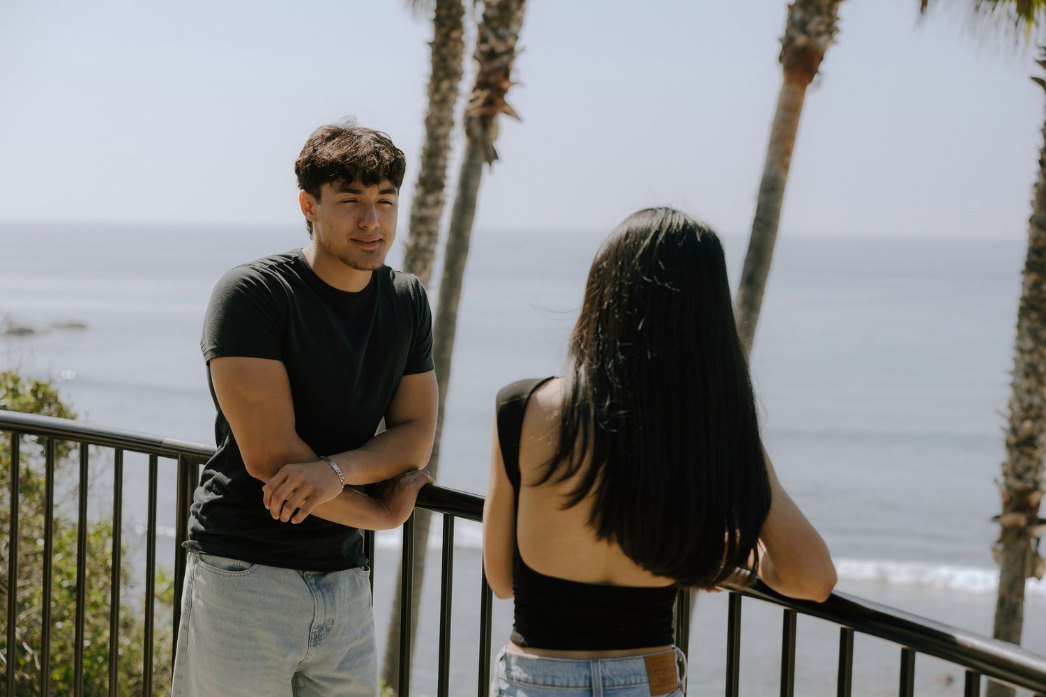 A young couple having a conversation on a seaside balcony with palm trees and ocean views in the background.