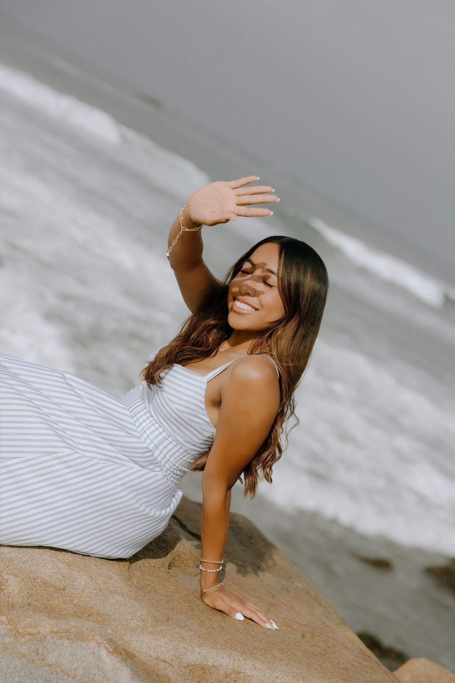 Smiling woman in striped dress sitting on rocks at beach, shielding eyes from sun with raised hand.