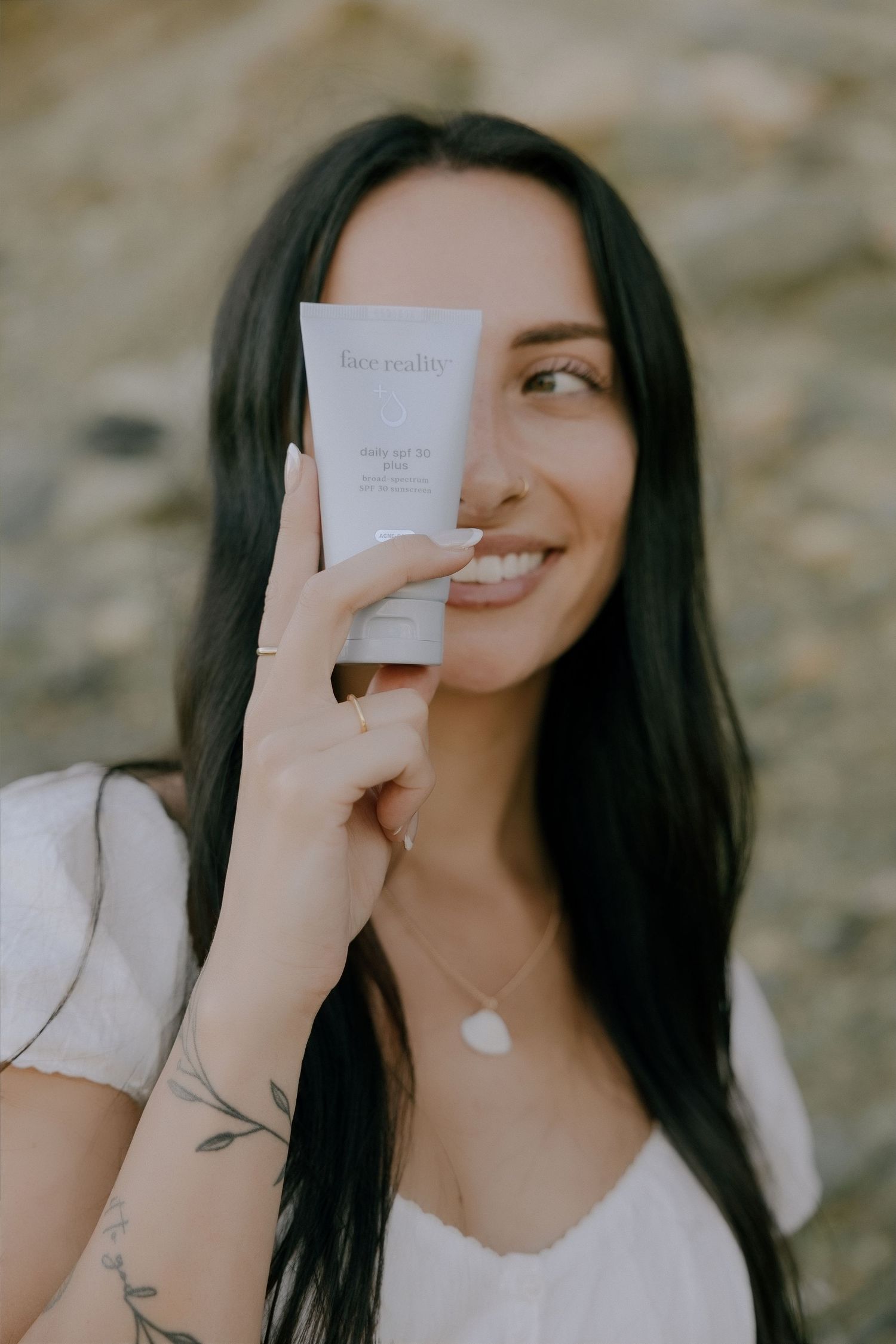 Woman smiling and holding Face Reality skincare product tube near her face outdoors.