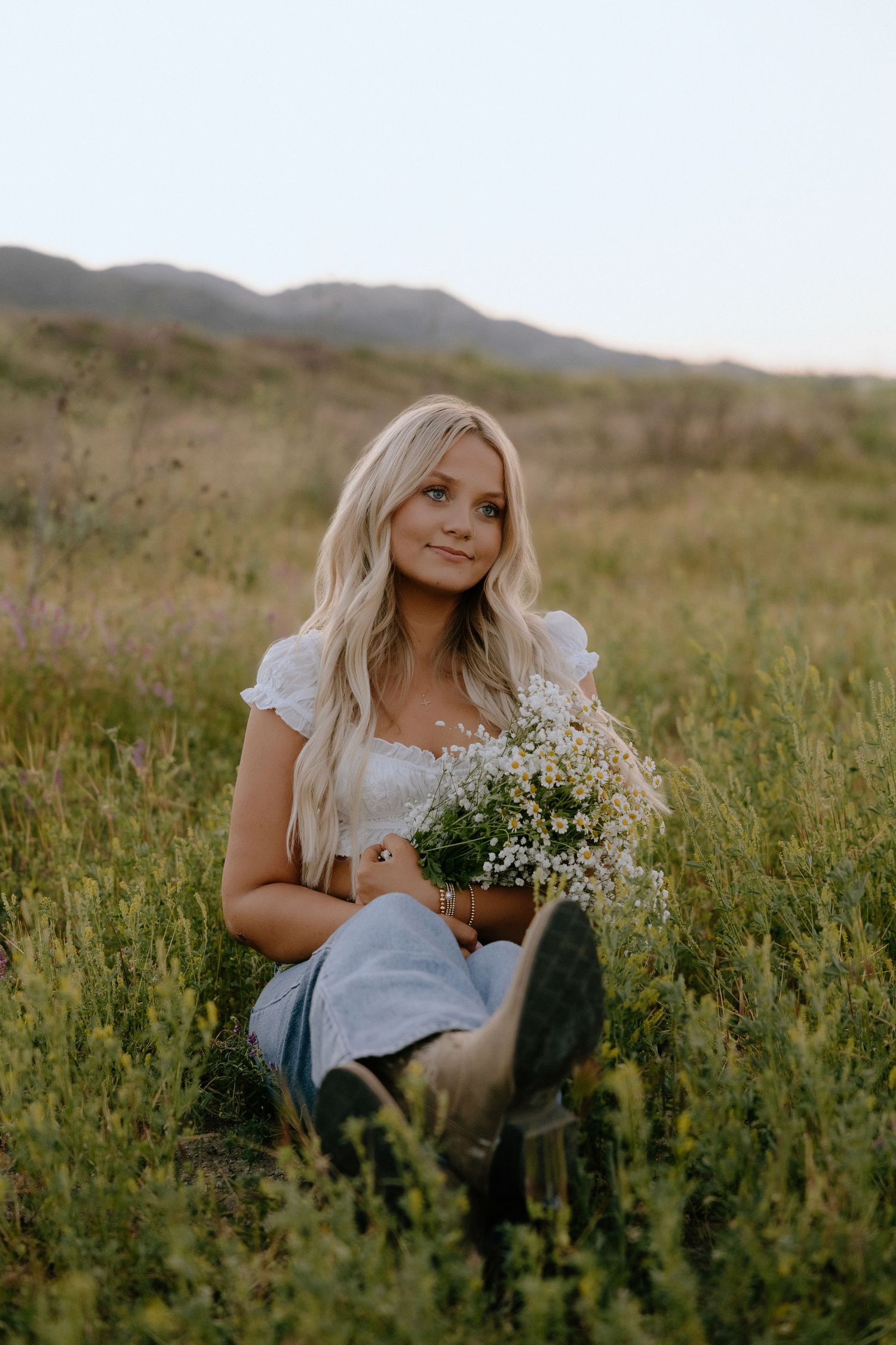 Young blonde woman sitting in a green field holding white wildflowers, wearing a white top and jeans with boots.