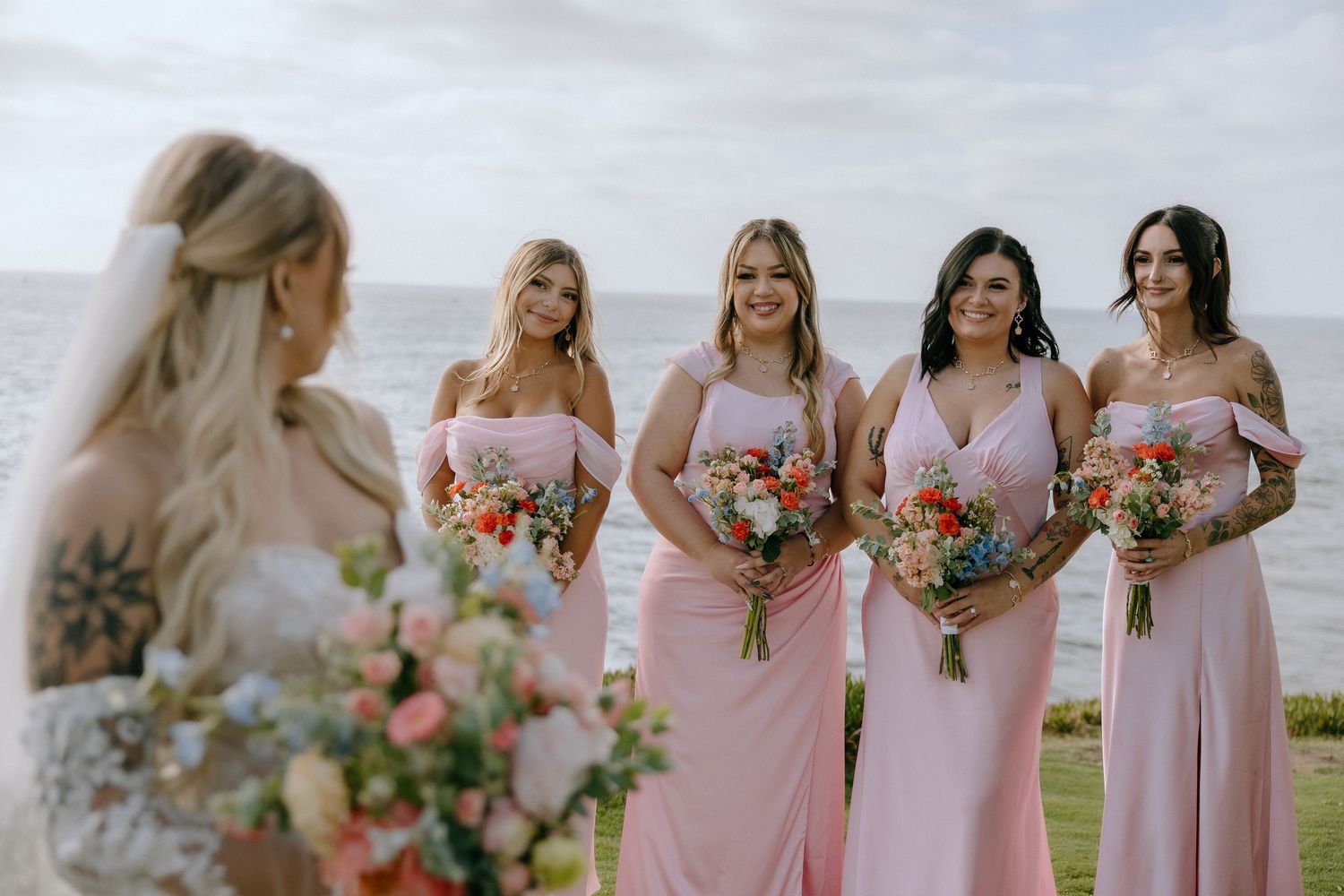 Bride faces three bridesmaids in pink off-shoulder gowns holding colorful bouquets by the ocean.