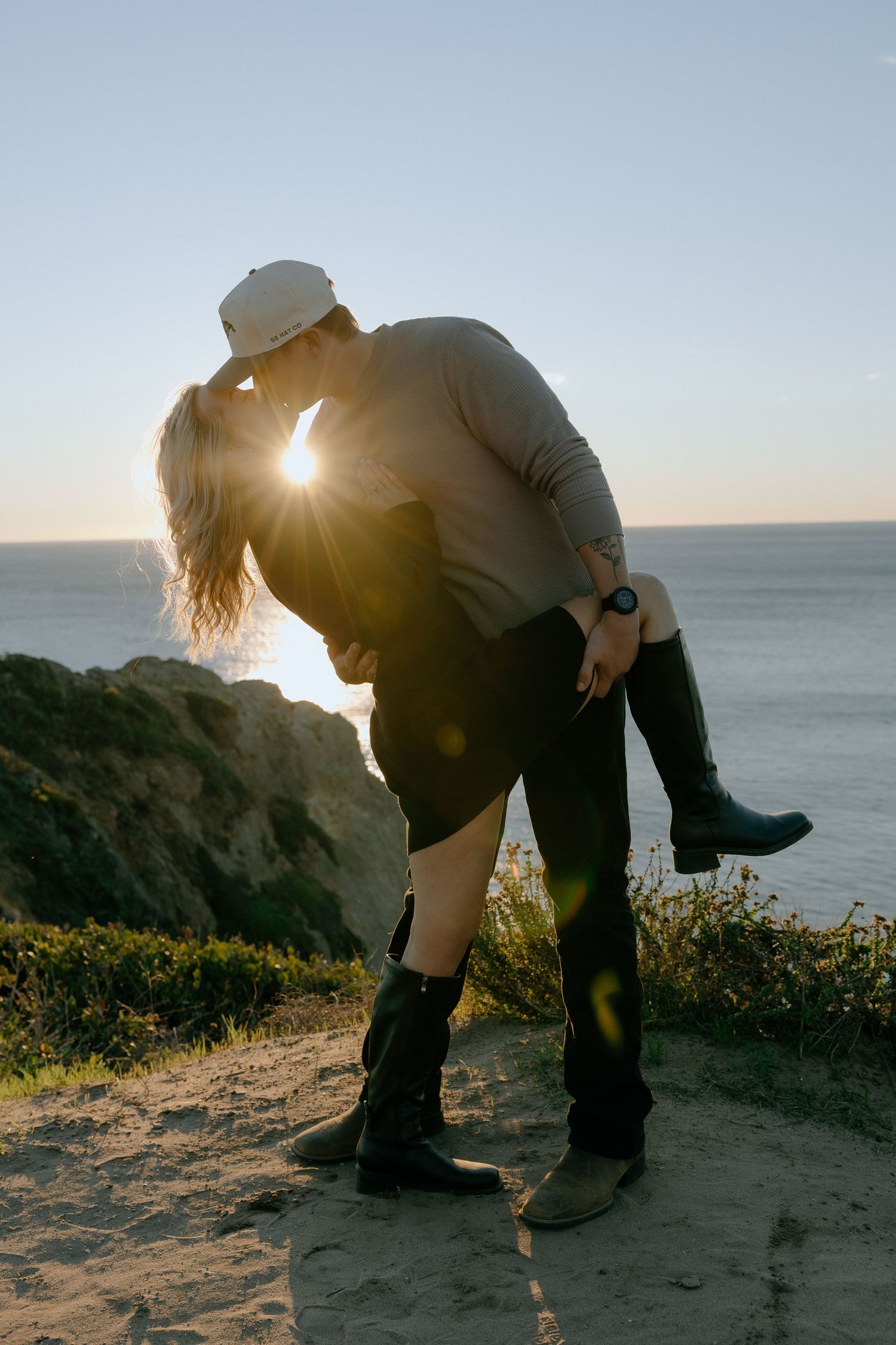 A romantic couple shares a sunset kiss on a coastal cliff overlooking the ocean.