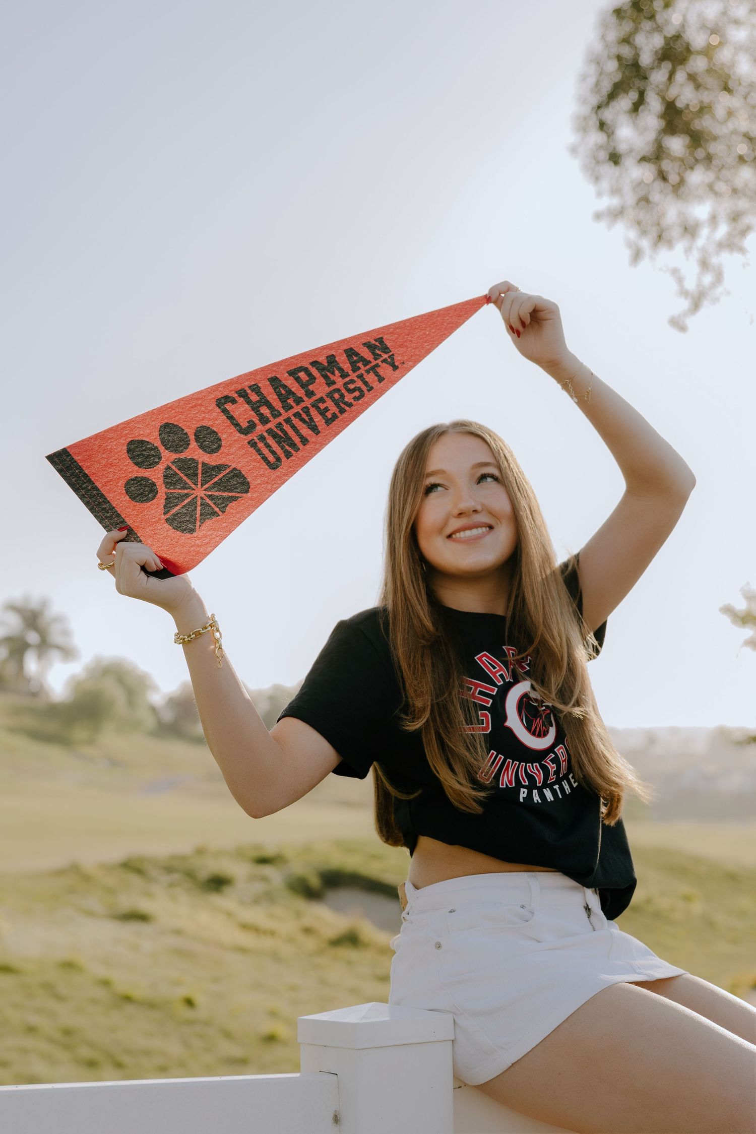 Young woman smiling and holding a Chapman University pennant flag outdoors on a sunny day.