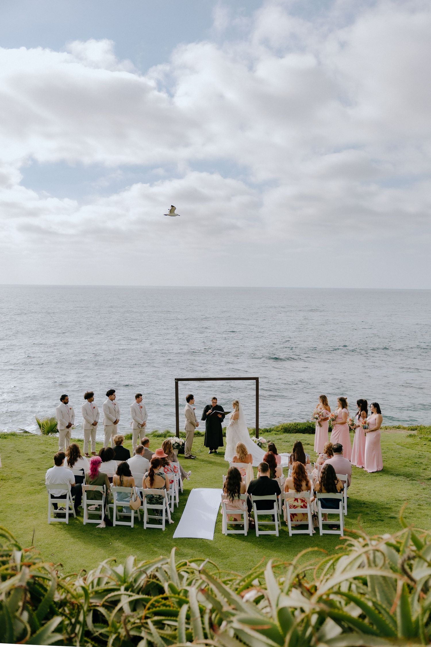 Outdoor oceanfront wedding ceremony on green lawn with white chairs, bridal party, and square arch under cloudy sky.
