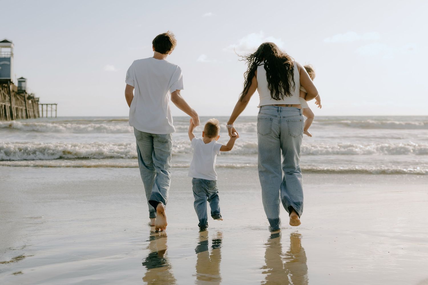 Family of three walking hand-in-hand on a sunny beach near a pier, viewed from behind.