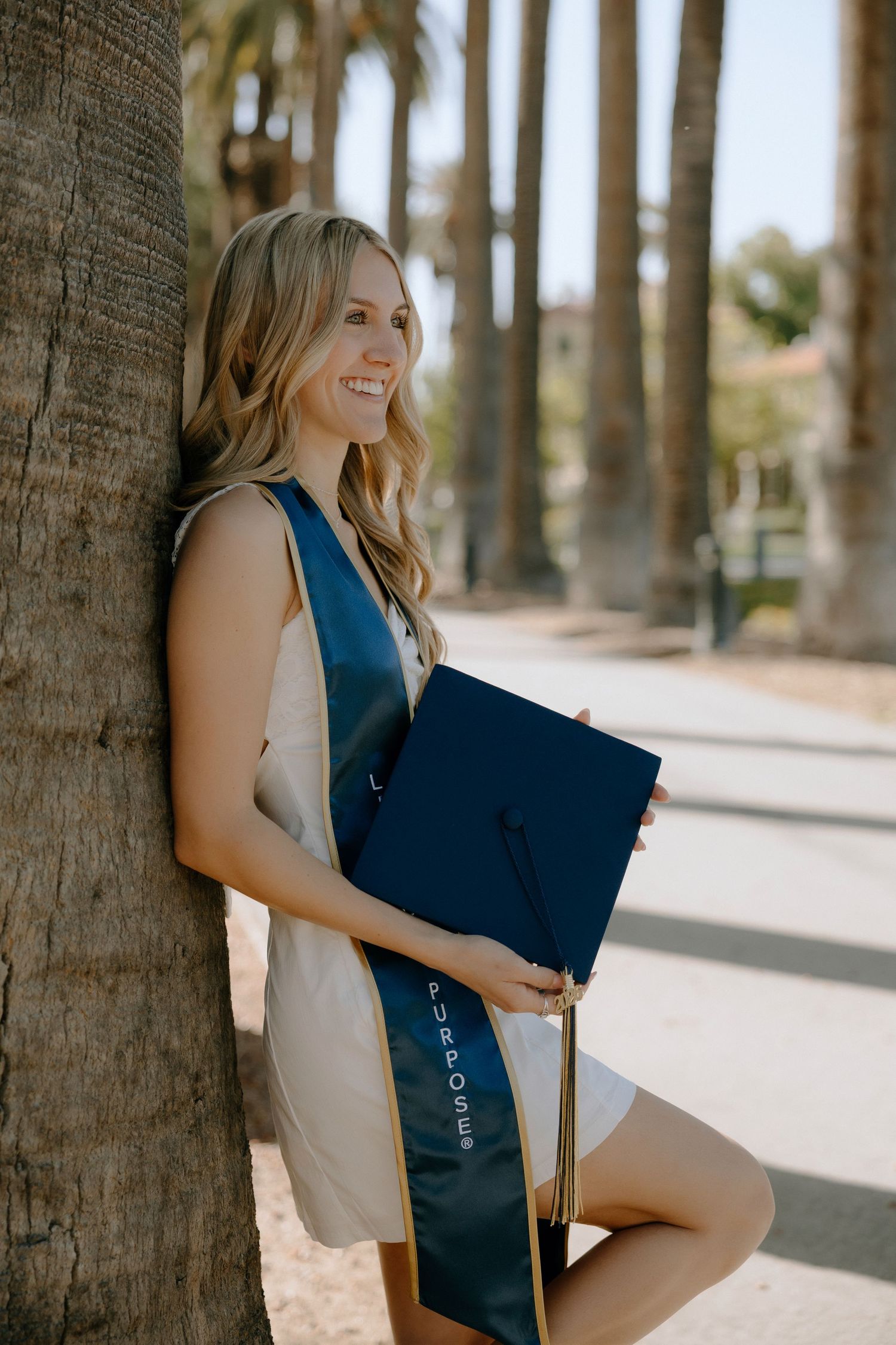 Smiling graduate in blue sash holds cap against palm tree, celebrating college graduation outdoors.